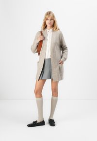 Junge Frau in beigem Cardigan, bluse in hellem Farbton, grauem Faltenrock, knielangen Socken, schwarzen Loafern und mit einer braunen Umhängetasche.