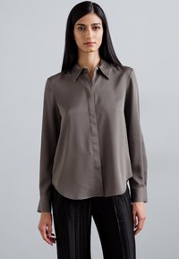 Long-sleeve, collared blouse in dark gray satin. Features a button-up front, smooth texture, and a rounded hem. Paired with pinstriped pants.