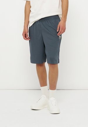 Mann iført mørkegrå knelengde shorts, hvite sokker, hvite joggesko og en lysebeige skjorte, stående mot en ensfarget hvit bakgrunn.
