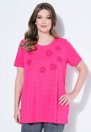 Frau mit welligem Haar, die große Creolenohrringe und ein leuchtend pinkes Hemd mit Blumenapplikationen trägt, steht mit einer Hand in der Hüfte.