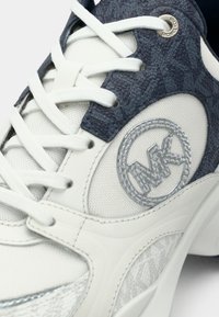 Λευκό αθλητικό παπούτσι με σκούρο μπλε υφασμάτινο πάνελ με υφή, ασημένιο έμβλημα λογότυπου και αντίθετα υλικά από δίχτυ. Τα κορδόνια είναι λευκά.