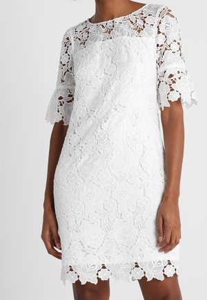 Žena v bílých květinových krajkových šatech délky po kolena s polovičními rukávy a vroubkovanými okraji na rukávech a lemu.