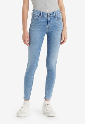 Femme portant un jean slim bleu clair, des baskets blanches et un débardeur gris côtelé, debout devant un fond blanc uni.
