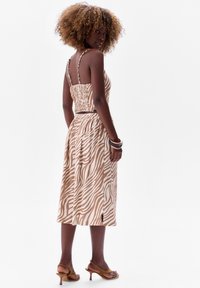 Sukienka w zebrowy wzór w odcieniach beżu i kremu, o średniej długości, z gumowanym tyłem, regulowanymi ramiączkami i ściągniętą talią. Stylizowana z brązowymi sandałami na obcasie.
