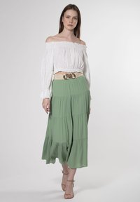 Blusa blanca off-the-shoulder de tejido texturizado, combinada con una falda verde de volantes y un cinturón con acentos dorados. La modelo lleva unos tacones neutrales.
