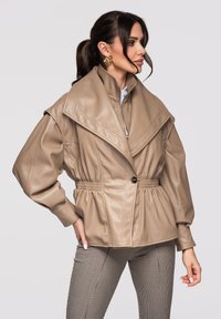 Mujer con cabello oscuro recogido en una coleta, lleva una chaqueta de cuero beige de gran tamaño con cuello ancho y cintura elástica, combinada con pantalones de cuadros.