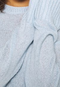 Hellblauer Strickpullover mit lockerem Muster und gerippten Bündchen. Die Textur wirkt weich und luftig und zeigt ein lässiges Design.
