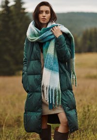 Žalias pūkinis paltas su siuvinėtu dizainu, ilgomis rankovėmis; per didelis multicolor šalikas, aptrauktas mėlynos ir žalios spalvų gradientu, su kutais.