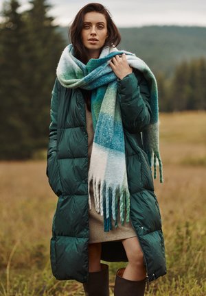 Grön puffjacka med quiltat mönster, långa ärmar; överdimensionerad flerfärgad scarf med gradientnyanser av blått och grönt, med fransar.