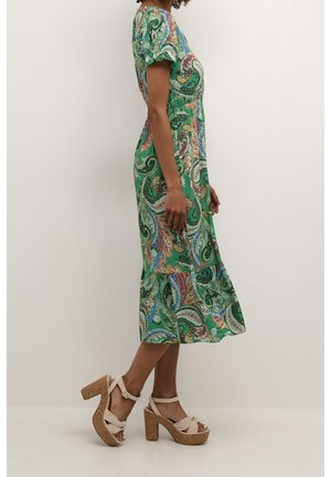 Femme portant une robe midi verte à motif cachemire et des sandales à talons hauts à plateforme beige, debout de profil contre un mur uni.
