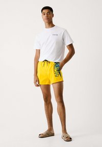Biela tričko s logom, žlté plavky s nápisom "RACING" v modrej farbe a hnedé šľapky; uvoľnený plážový outfit.