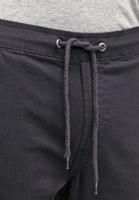 Černé kalhoty na šňůrku s pletenou stahovací šňůrkou, s hladkou texturou a kovovými průchodkami v pase. Nad kalhotami je vidět šedý materiál.