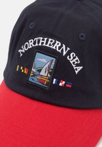 Căciulă bleumarin și roșie, din bumbac, cu un design brodat "Marea Nordică" și temă marinară, cu steaguri și bărci cu pânze.