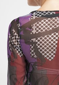 Blusa trasparente viola e nera con pois e motivi astratti, dotata di scollatura rotonda e maniche lunghe. Tessuto morbido e leggero.