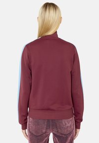 Burgundská mikina s modrými bočními pruhy, s vysokým límcem a ribovanými manžetami. Hladká bavlněná tkanina a volný střih.