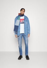 Chaqueta de mezclilla, azul claro con bolsillos, superpuesta sobre una camiseta gráfica gris. Combinada con jeans azules desgastados y una bufanda de colores variados con flecos.