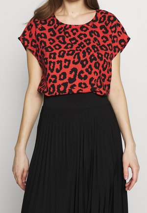 Woman wearing a red short-sleeve blouse with black leopard print and a high-waisted black pleated skirt against a plain background.