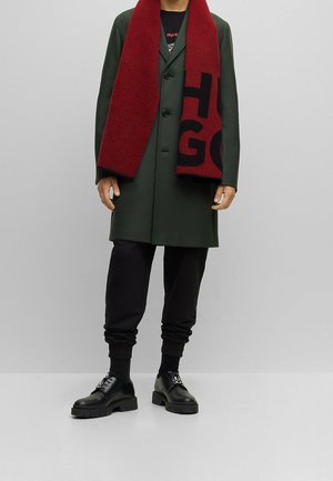 Homme portant un pardessus vert foncé, une écharpe rouge et noire avec de grandes lettres, un pantalon noir et des chaussures noires, sur fond gris uni.