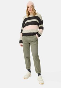 Vrouw in een roze beanie, gestreepte fuzzy trui, olijfgroene broek, grijze sokken en witte sneakers, staand met één hand in de zak.
