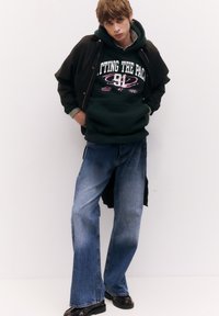 Felpa con cappuccio verde scuro con grafica bianca e rosa, indossata sotto una giacca nera. Jeans azzurri a gamba larga e scarpe nere completano il look.