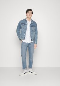 Chaqueta de denim en azul claro con botones plateados y bolsillos en el pecho, combinada con una camisa blanca y jeans azules claros. Zapatillas blancas con detalles negros.