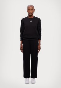 Zwarte sweatshirt met EA7-logo, gecombineerd met zwarte sweatpants met zijzakken en wit logo. Witte sneakers maken de outfit af.