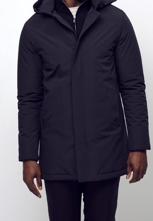 Mężczyzna w czarnej zimowej kurtce z kapturem, nałożonej na czarną zasuwaną kurtkę i białą koszulę, stojący na tle jednolitego jasnego tła.