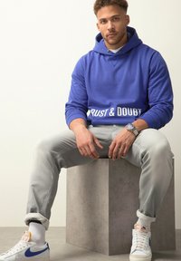 Sudadera de algodón azul con el texto "CONFIANZA Y DUDAS", jeans ajustados grises y zapatillas blancas con acentos azules. Modelo sentado sobre un pedestal de concreto.