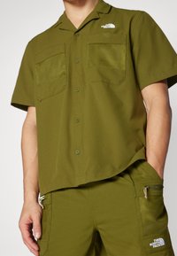 Oliwkowa koszula z krótkim rękawem, ozdobiona dwoma siateczkowymi kieszeniami, zapinana na guziki z przodu, o elastycznym kroju zapewniającym wygodę i styl.