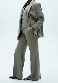 Jasnoszary garnitur skrojony z fakturowanej tkaniny, z szerokimi spodniami i dopasowaną marynarką z dwoma guzikami oraz jedną kieszenią na piersi.