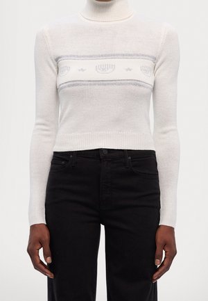 Personne portant un pull court à col roulé blanc avec un motif gris de menorah et d'étoile et un pantalon noir taille haute, debout avec les bras détendus.