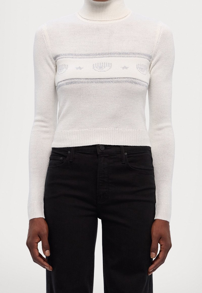 Personne portant un pull court à col roulé blanc avec un motif gris de menorah et d'étoile et un pantalon noir taille haute, debout avec les bras détendus.