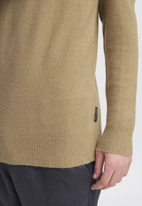 Beige strikkegenser med ribbestrikkede mansjetter og nederkant. Har et sidetagg. Myk tekstur, tett passform og lange ermer.