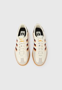 Beli tekstilni supergi z rjavimi semišnimi poudarki, črnimi in rdečimi črtami, teksturirano gumijasto podplato in okroglimi vezalkami.