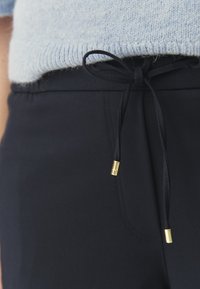 Gros plan sur un pantalon noir avec un cordon de serrage noué orné d'embouts métalliques dorés, associé à un haut en tricot bleu clair.