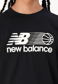 Črna bombažna majica s kratkimi rokavi, ki ima bel grafik z napisom "NB" in "new balance", skupaj z obliko koša za košarko. Okrogli izrez, mehak material.