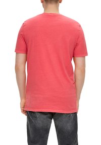 Korallenfarbiges T-Shirt mit kurzen Ärmeln, Rundhalsausschnitt, weiche Textur, mit subtilen Branding-Details nahe dem Saum, getragen mit dunklen Jeans.