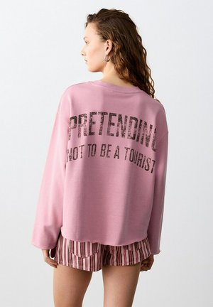 Junge Frau mit lockigem Haar, die einen rosa Pullover mit der Aufschrift "So tun, als wäre ich kein Tourist" und gestreifte Shorts trägt, seitlich zugewandt.