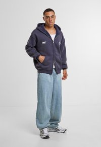 Sudadera oscura de color marino con cremallera y bolsillo frontal, con acentos de logotipo blanco. Combinada con unos vaqueros de pierna ancha de color azul claro y zapatillas de running plateadas.