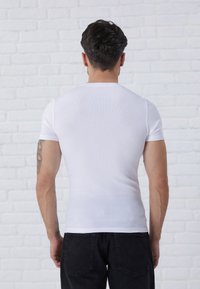 Weißes, geripptes T-Shirt mit kurzen Ärmeln, taillierter Passform, glatter Textur und rundem Halsausschnitt, kombiniert mit dunklen Jeans vor einem weißen Ziegelhintergrund.