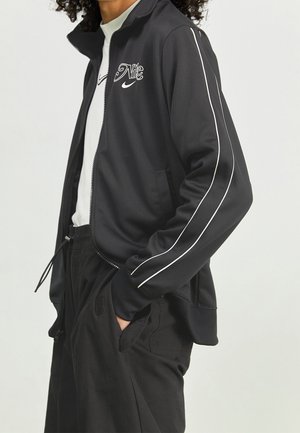 Personne portant une veste zippée noire Nike avec des liserés blancs sur les manches et le logo Nike, associée à un pantalon noir et un t-shirt blanc Nike.