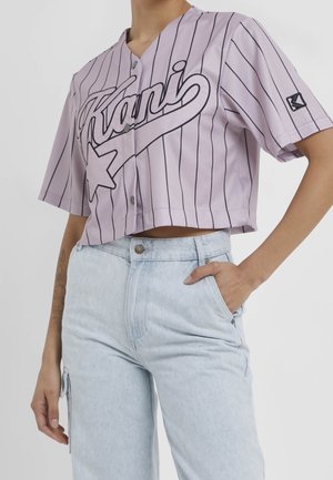 Woman wearing a cropped lavender baseball shirt with black pinstripes and logo, paired with light blue high-waisted jeans with side pocket.