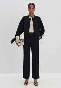 Zwarte jas met knopen, losse pasvorm; witte crop top; op maat gemaakte zwarte broek; met een zwart en beige handtas; platte sandalen.