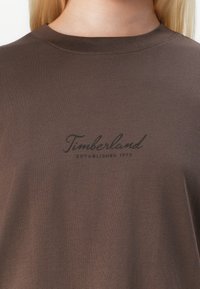 Tricou din bumbac maro cu guler rotund și logo-ul Timberland în negru, având inscripția "ESTABLISHED 1973" dedesubt. Textură netedă.