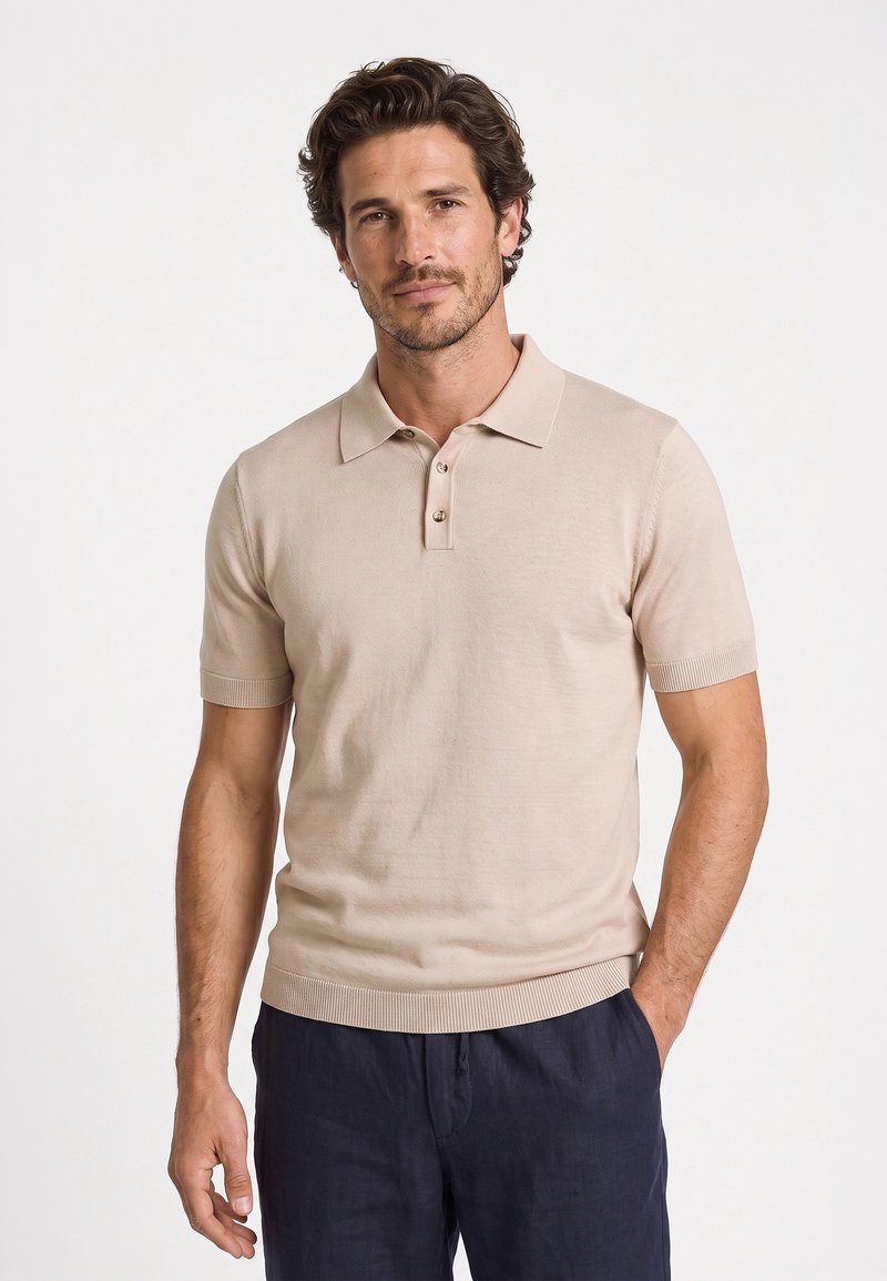 Mand iført beige kortærmet polo og mørke bukser står med den ene hånd i lommen mod en ensfarvet hvid baggrund.
