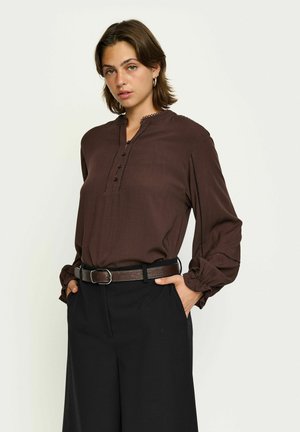 Chemise marron à boutonnière avec un col mandarin, en tissu texturé, et des poignets de manches bouffantes, assortie à un pantalon noir taille haute et une ceinture marron.