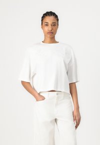 Witte geschikte T-shirt met korte mouwen, versierd met bloemenborduursel over de borst. Gekleed met witte high-waisted broek.