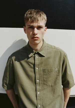 Olive green short-sleeve button-up shirt with a chest pocket and a small embroidered logo. Fabric has a soft texture and subtle sheen.