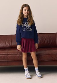 Felpa blu navy con logo grafico, gonna plissettata color bordeaux e sneakers bianche con accenti blu. La modella è in piedi davanti a un divano in pelle marrone.
