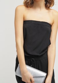 Jumpsuit noir sans bretelles avec un dessus élastiqué, associé à une pochette à paillettes argentées. La pochette a un design rectangulaire et élégant.